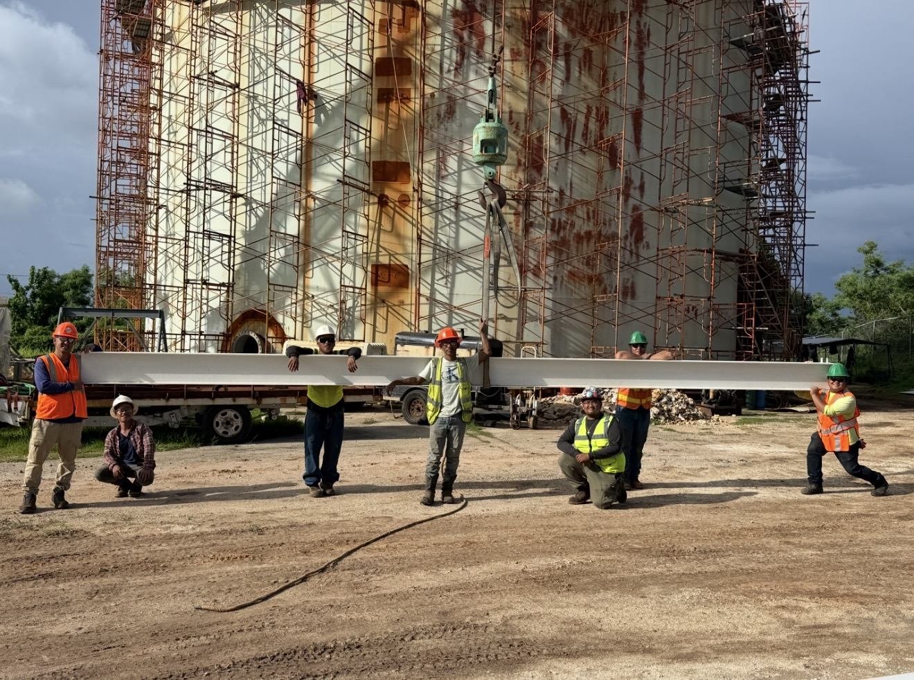 RDE crew in hard hats and safety vests lined up along a steel I-beam on the crane hook at the Malojloj tank